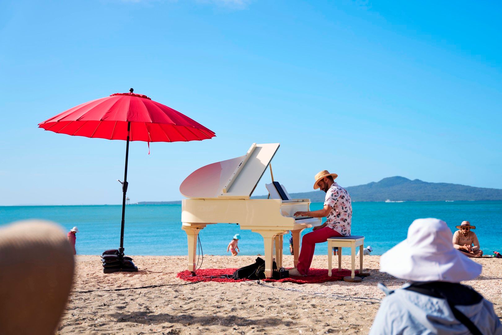 A side shot of a person seated at the piano on a beach, in a wide straw hat and colourful shirt. The ocean and edge of Rangitoto are visible behind it, and a bright red sun umbrella.